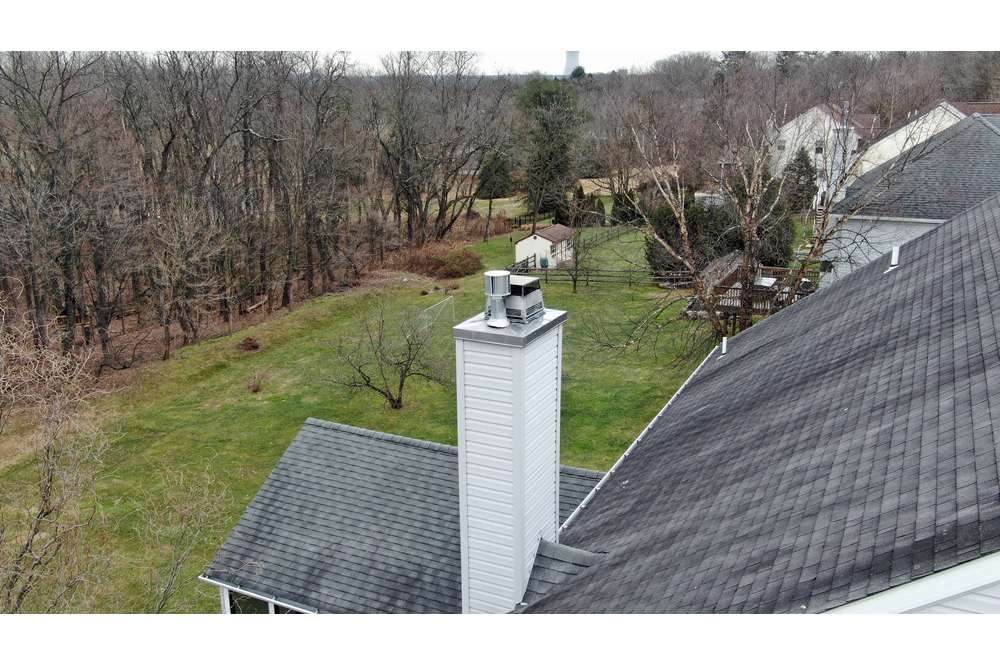 A gray roofed house with a prominent white chimney surrounded by trees and a green yard in a suburban setting, serviced by the best chimney company Long Island has to offer.